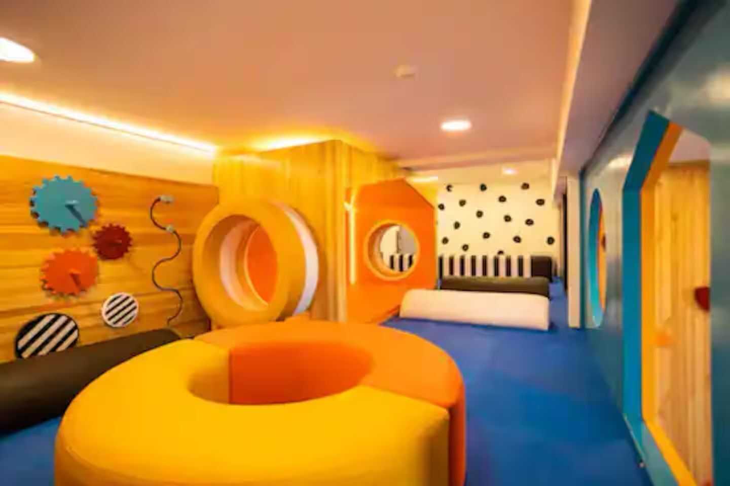 A room with a blue carpet, orange and yellow play structures, and a wall with a clock and circular decorations.