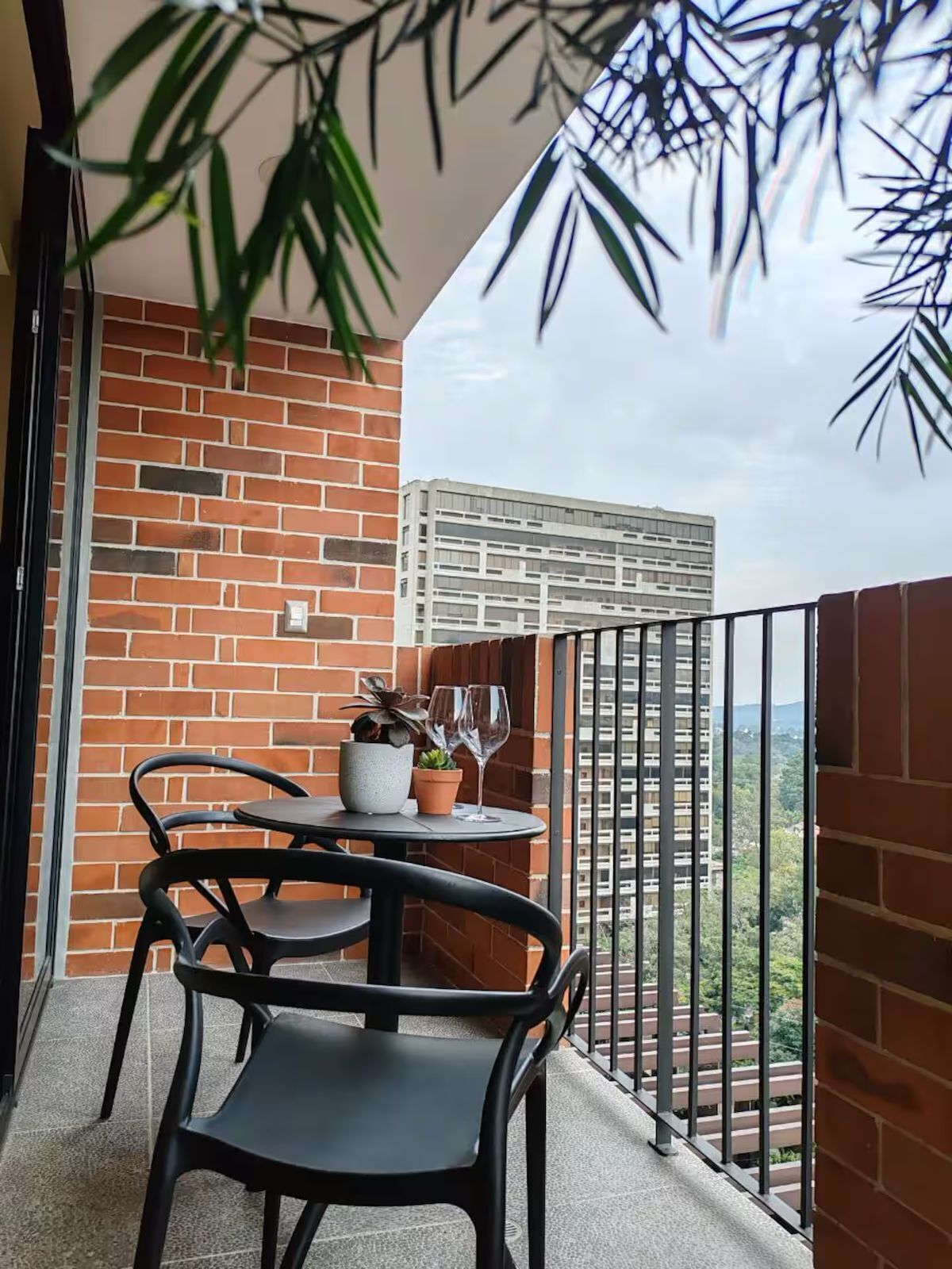 A balcony with a brick wall, a small table with two chairs, a potted plant, and a wine glass.