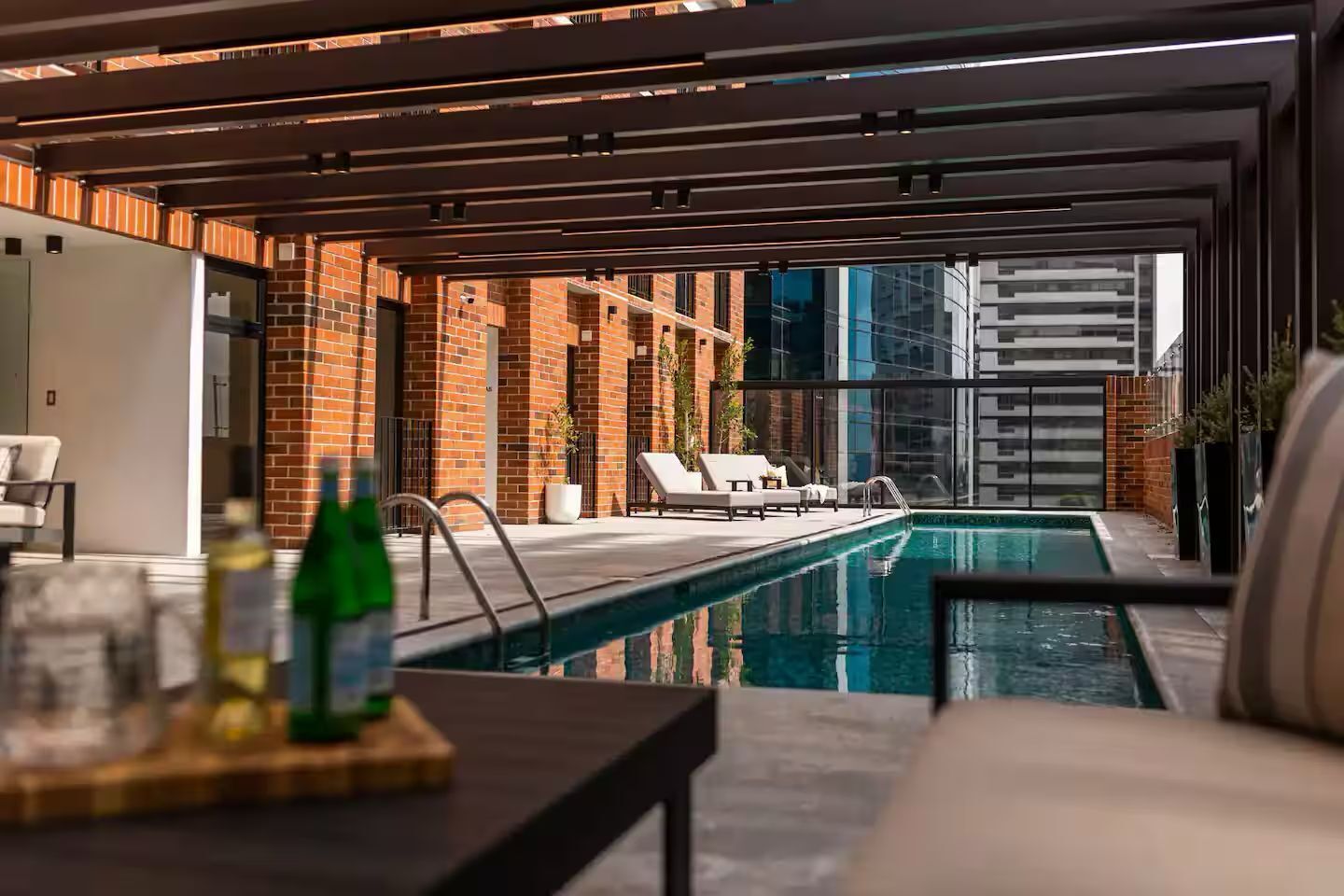 Un área de piscina moderna al aire libre con sillones de descanso, un muro de ladrillo y un edificio de cristal al fondo.
