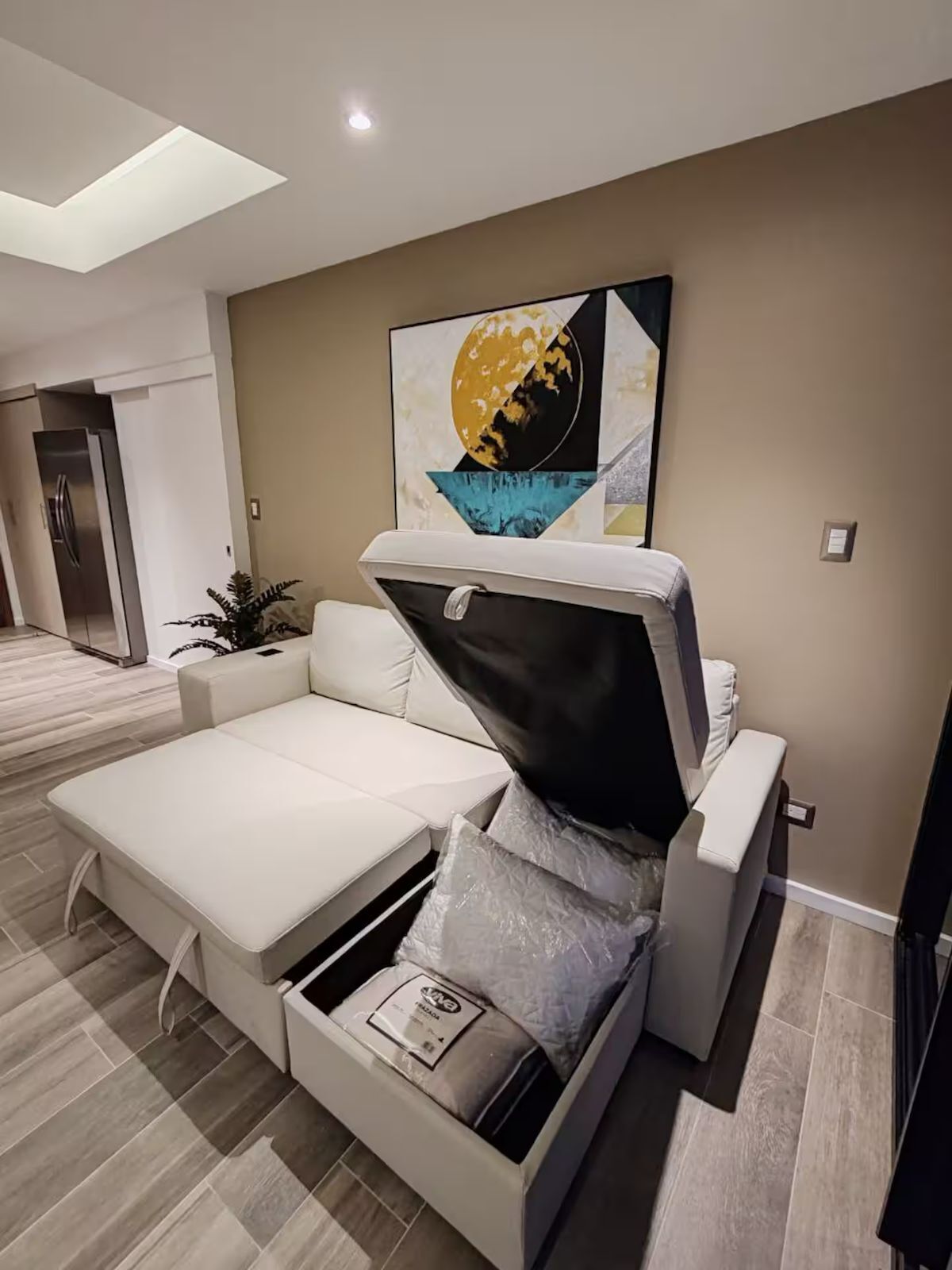 A modern living room with a sofa bed, a painting on the wall, and a kitchen in the background.
