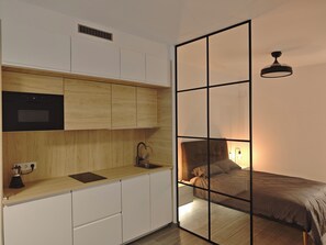 A compact bedroom with a bed, bedside table, and a small kitchenette area with a microwave, sink, and storage cabinets.