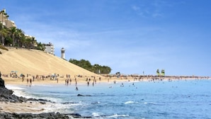 Uma praia com areal, um farol e uma multidão de pessoas.