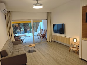 A modern living room with a sofa, a TV mounted on the wall, and a sliding glass door leading to an outdoor area with plants.