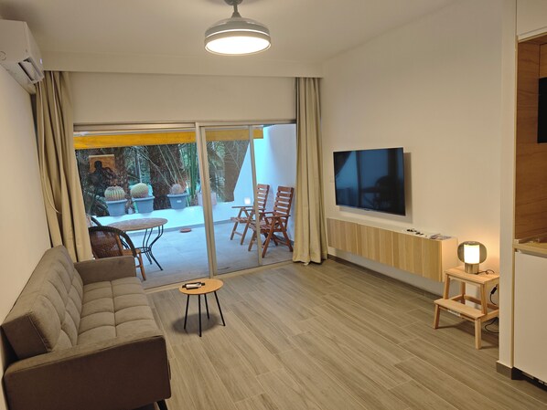 A modern living room with a sofa, a TV mounted on the wall, and a sliding glass door leading to an outdoor area with plants.