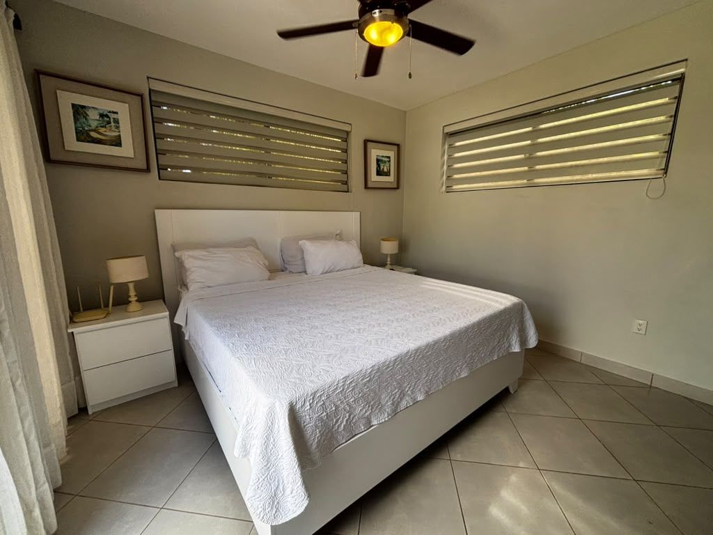 A bedroom with a bed, nightstands, a ceiling fan, and wall-mounted blinds.