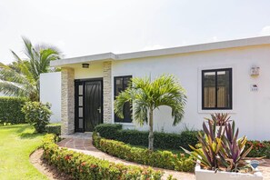 Una casa moderna de dos pisos con fachada de piedra, una puerta negra y un pequeño jardín con diversas plantas y una palmera.