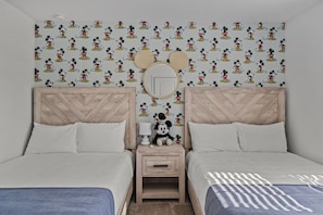 Un dormitori amb dos llits, una nit del taulell amb una figura de peluix del Micky Mouse, i una paret amb paper pintat del Micky Mouse.