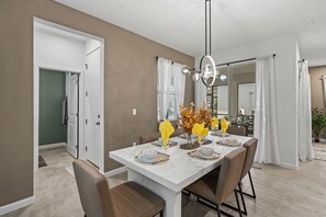 A dining area with a white table set for four, a chandelier, and a view of a kitchen and dining area through a glass door. - Brand New | 6BR Villa Pool & Spa,Loft near Disney 2380 (Kissimmee)