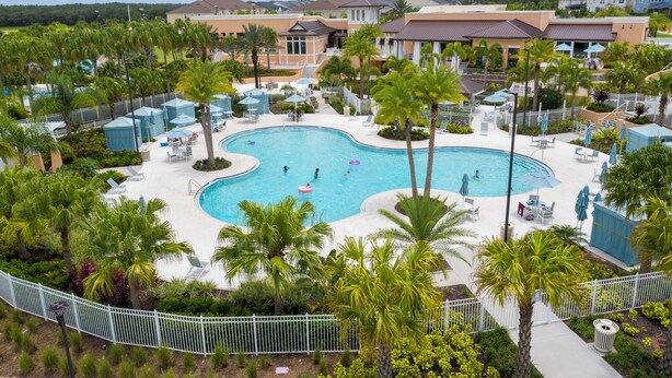 A large outdoor swimming pool surrounded by palm trees and lounge chairs, with a view of residential buildings in the background. - Brand New | 6BR Villa Pool & Spa,Loft near Disney 2380 (Kissimmee)