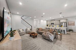 A modern living room with a sofa, coffee table, and a dining area with chairs and a kitchen in the background. - Brand New | 6BR Villa Pool & Spa,Loft near Disney 2380 (Kissimmee)