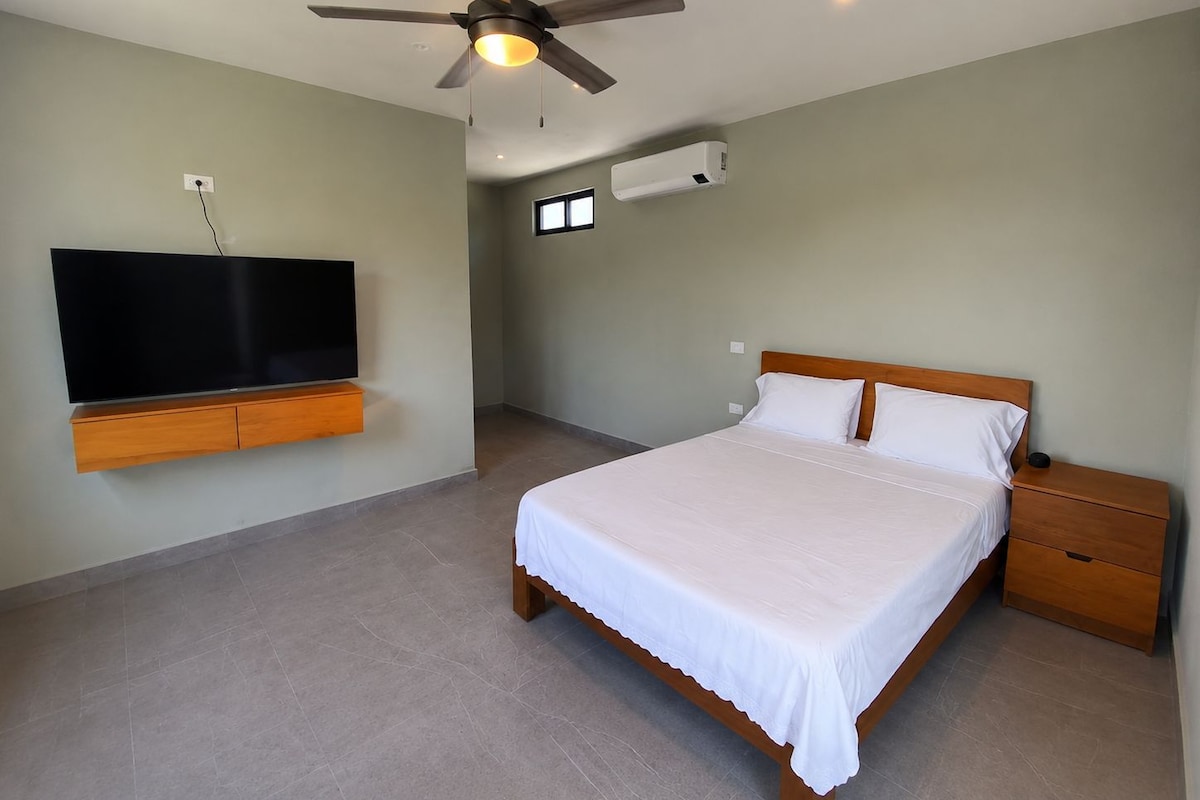 A bedroom with a bed, a TV mounted on the wall, a ceiling fan, and an air conditioning unit.