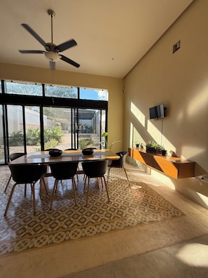 Un comedor con una mesa, sillas, un ventilador de techo y vistas amplias a través de ventanas grandes.