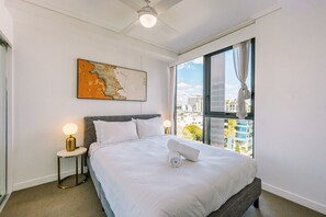 A hotel room with a large bed, a view of the city, a ceiling fan, and a painting on the wall. - One Bedroom Urban Solace Near CBD (Fortitude Valley)