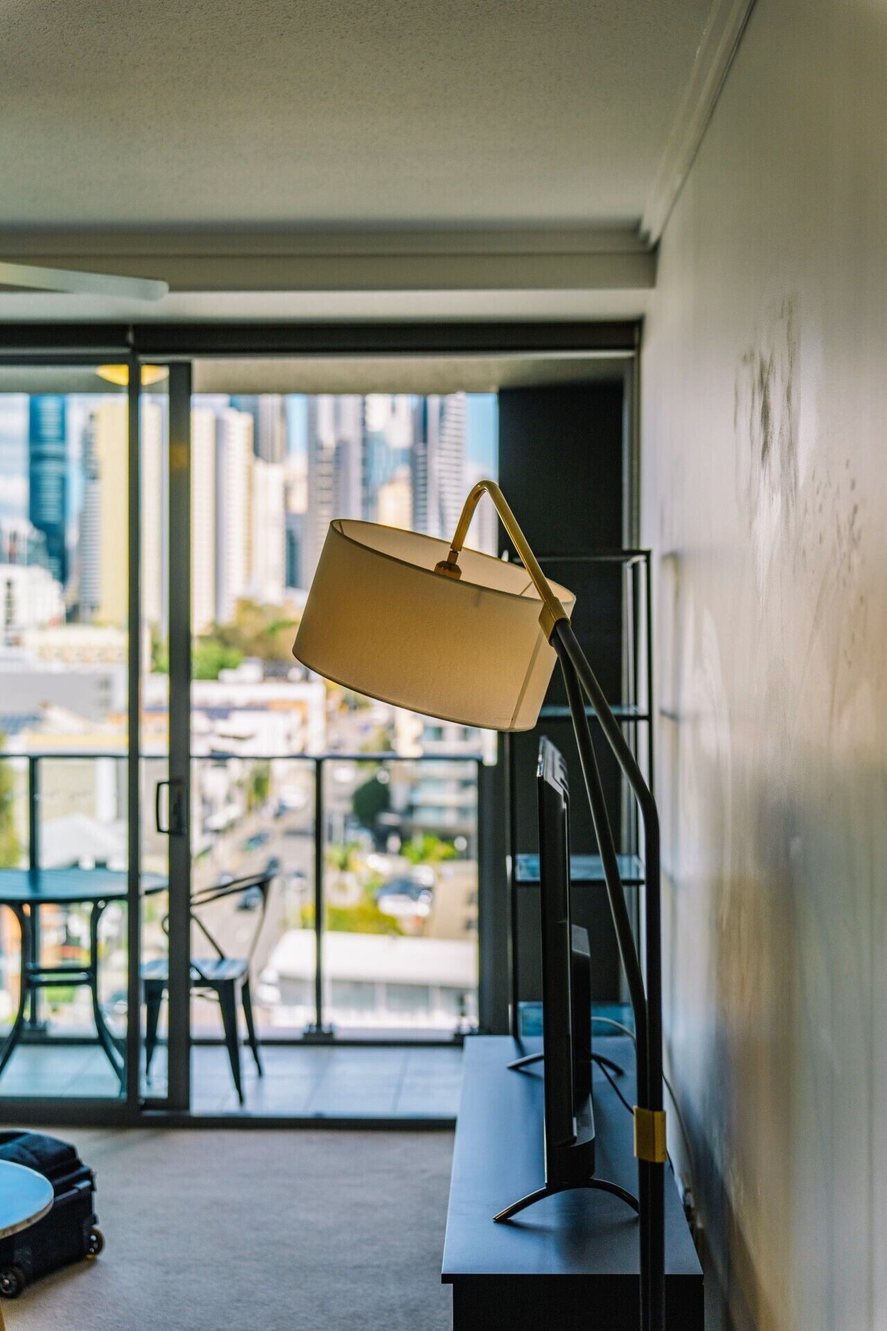 A room with a desk, a lamp, and a view of a cityscape through large windows.