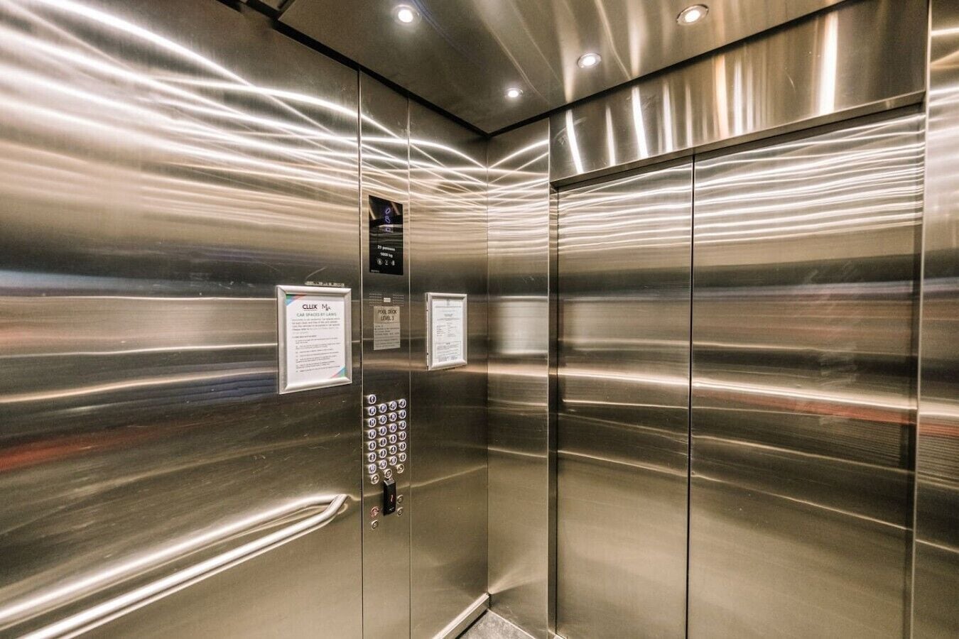 Elevatorinteriør med vægge i rustfrit stål, et kontrolpanel med knapper og informationsskilte.