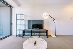 A modern living room with a flat-screen TV, a standing lamp, a glass shelf unit, and a remote control on a marble table. - One Bedroom Urban Solace Near CBD (Fortitude Valley)