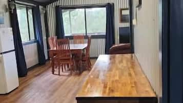 A room with a wooden table and chairs, a refrigerator, and large windows.