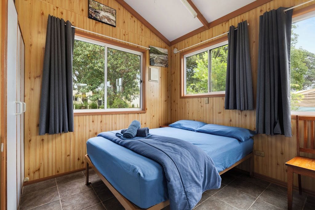 Een kamer met houten wanden, twee bedden met blauw beddengoed, grote ramen met gordijnen en uitzicht op groen buiten.