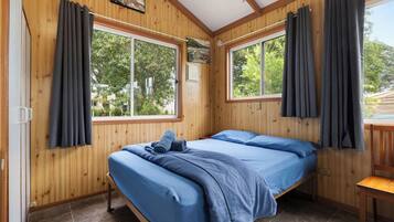 Een kamer met houten wanden, twee bedden met blauw beddengoed, grote ramen met gordijnen en uitzicht op groen buiten.
