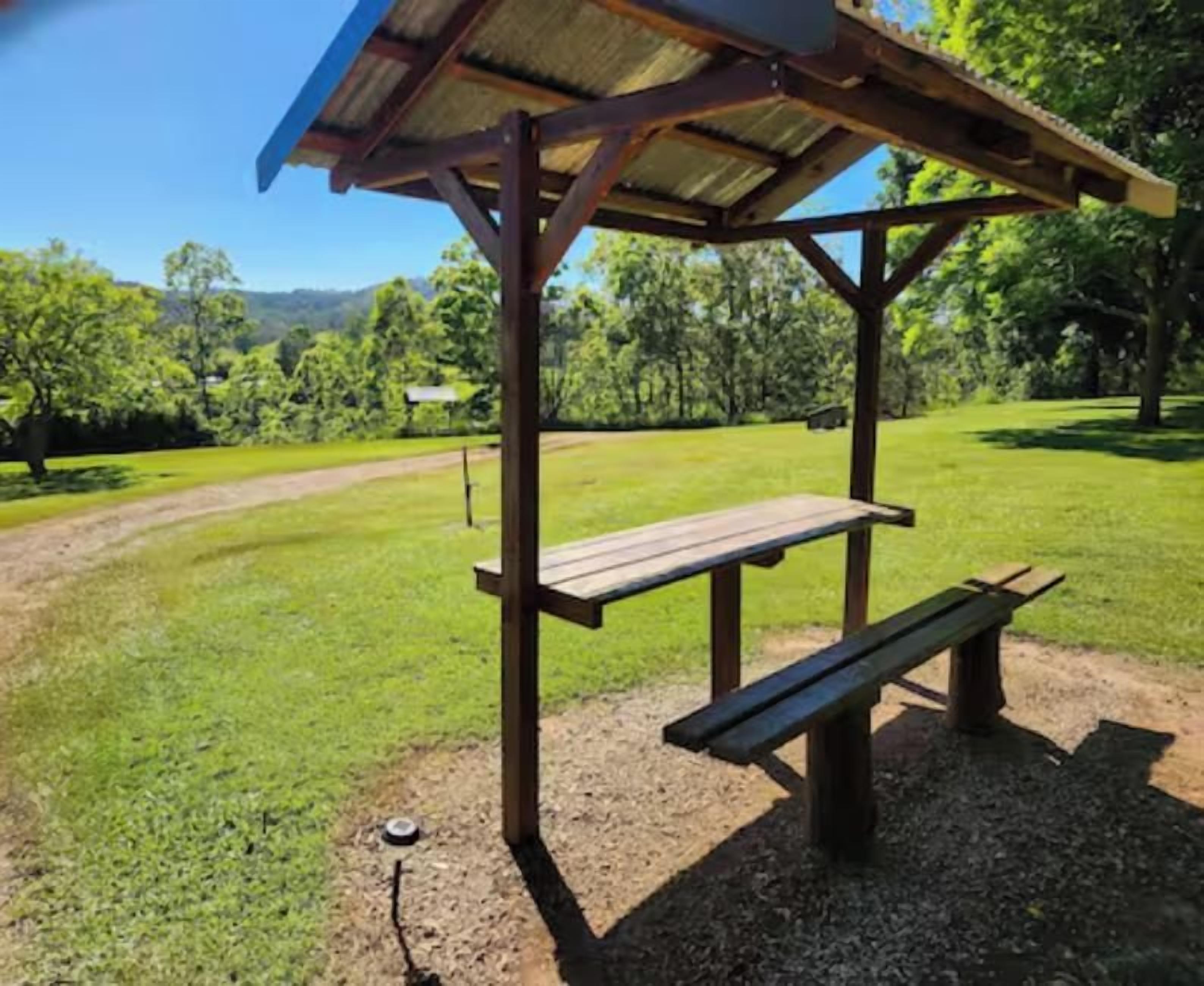 A wooden picnic shelter with benches, set on a grassy area with a dirt path and trees in the background.
