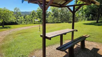 A wooden picnic shelter with benches, set on a grassy area with a dirt path and trees in the background.