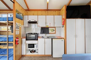 A compact kitchen with a microwave, toaster, and small refrigerator. There is a bunk bed with blue bedding to the left.