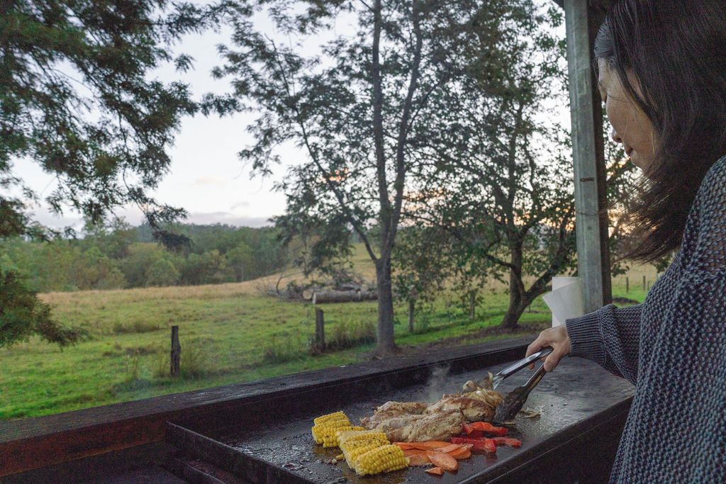 A person grilling food outdoors with a view of a grassy field and trees in the background.
