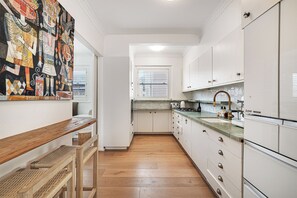 Une cuisine avec des meubles blancs, un banc en bois et un mur orné d’un tableau abstrait aux couleurs vives.