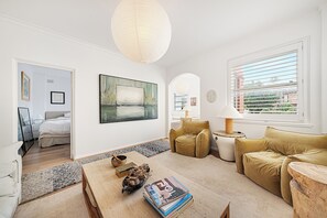A modern living room with a sofa, a coffee table with books, a painting, and a window with blinds. - Casa Franka - Art Deco Beachfront Apartment in North Bondi (Bondi Beach)
