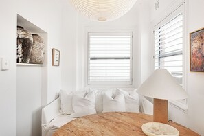 Une pièce avec une table ronde en bois, un canapé blanc avec des coussins, un grand vase, un tableau encadré et une fenêtre avec des stores.