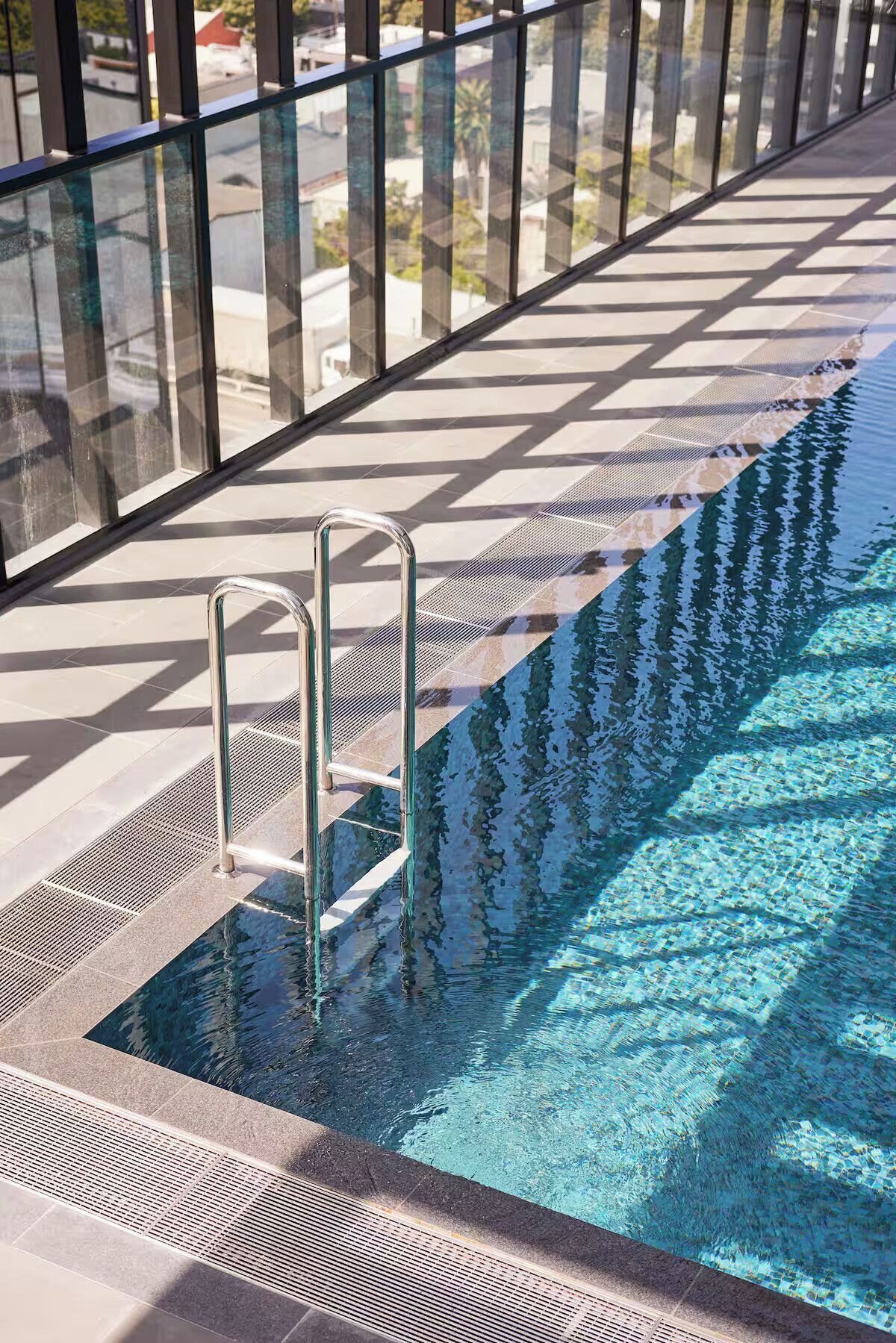 Piscina com escada de metal, rodeada por guarda-corpo de vidro e um deck revestido com azulejos.