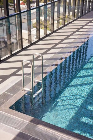En swimmingpool med en metalstige, omgivet af gelænder i glas og en flisebelagt terrasse.