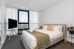 A hotel room with a bed, a TV on a stand, a window with curtains, and a view of the city. - Skyline & Bay Views Stylish 3BR Southbank Retreat (Southbank)