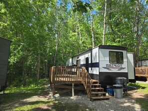 A recreational vehicle (RV) with a wooden deck attached, surrounded by trees.