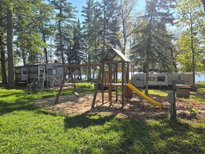 A playground with swings and a slide, surrounded by trees and RVs.