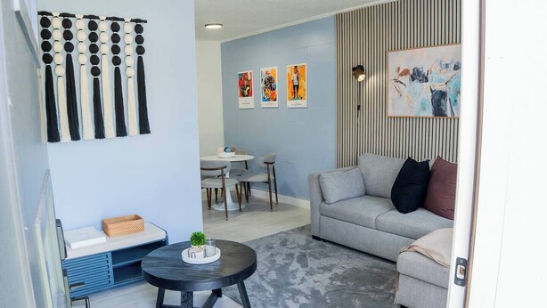 Ein modernes Wohnzimmer mit einem grauen Sofa, einem runden schwarzen Couchtisch und einem Essbereich mit weißem Tisch und Stühlen.