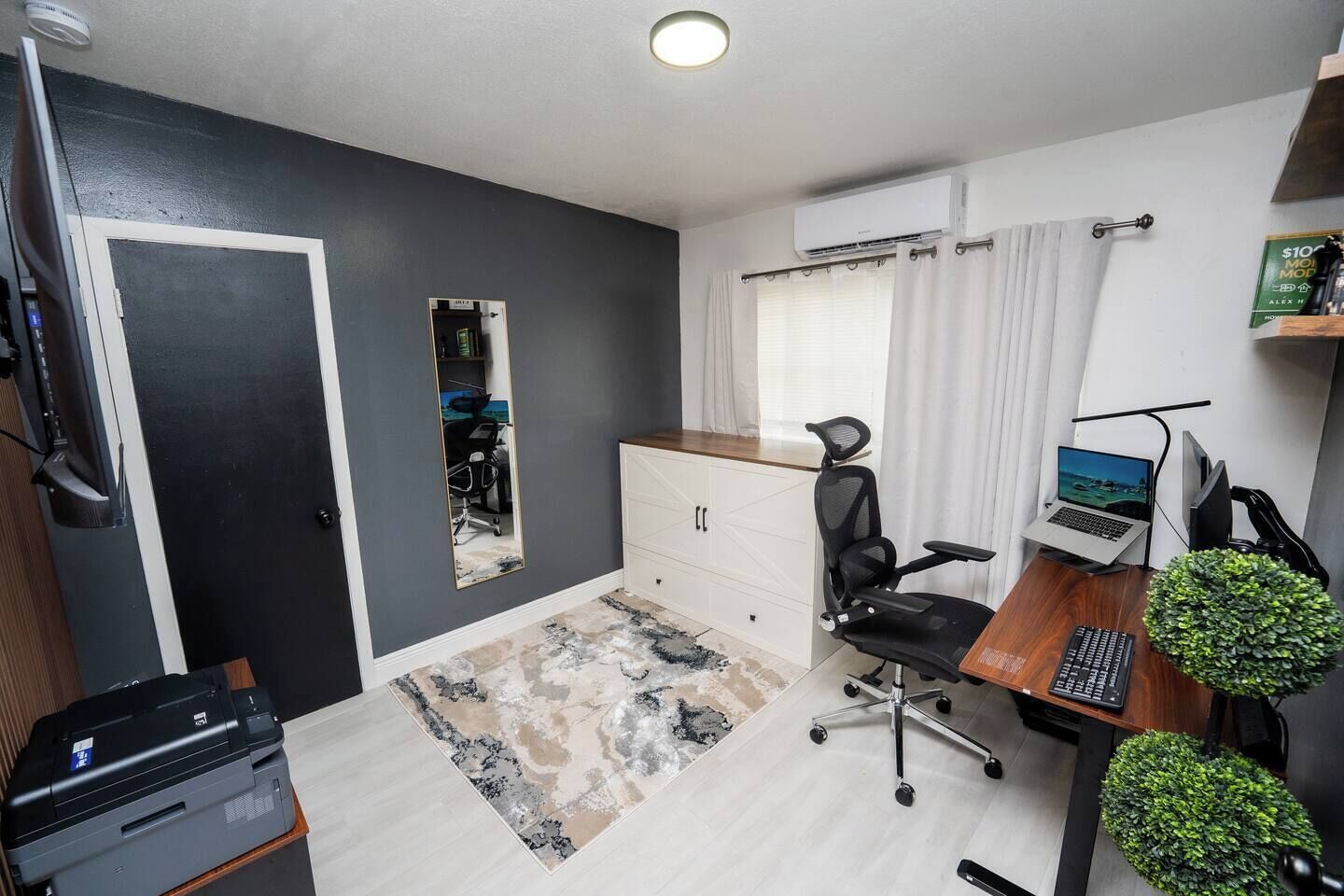 Ein Zimmer mit Schreibtisch, Stuhl, Computer, Drucker und einer kleinen Pflanze.
