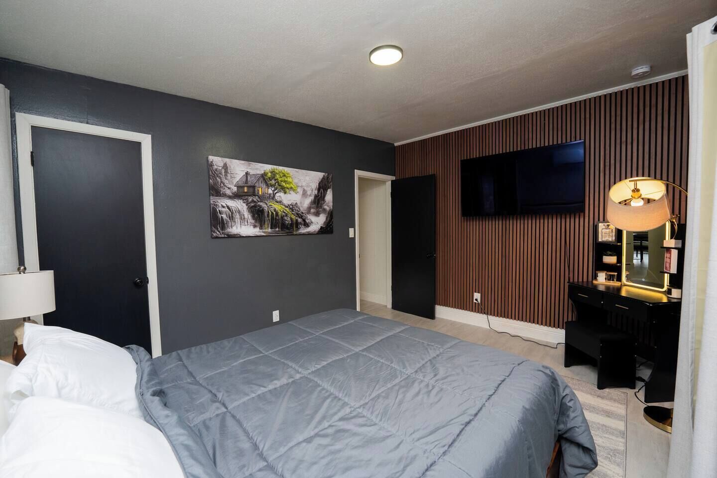 Een slaapkamer met een bed, een aan de muur bevestigde televisie, een bureau met een lamp en een schilderij.