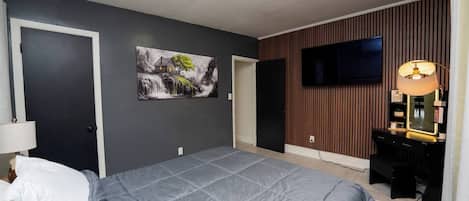 Une chambre à coucher avec un lit, un téléviseur fixé au mur, un bureau avec une lampe et un tableau.