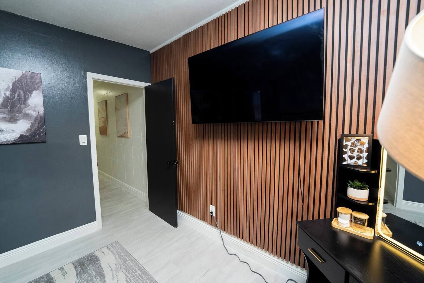 A modern living room with a flat-screen TV mounted on a wooden wall, a door leading to another room, and a small table with decorative items.