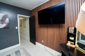 A modern living room with a flat-screen TV mounted on a wooden wall, a door leading to another room, and a small table with decorative items. - Nomad Haven in Downtown: Recharge, Work & Explore (Orlando)