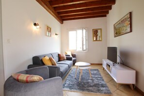 Sala de estar com sofá cinza, uma mesinha branca, TV em um móvel branco e janela com cortinas.