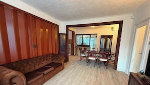Una sala de estar con un sofá marrón, paneles de madera en las paredes, una zona de comedor con una mesa y sillas, y una cocina visible a través de un vano abierto.