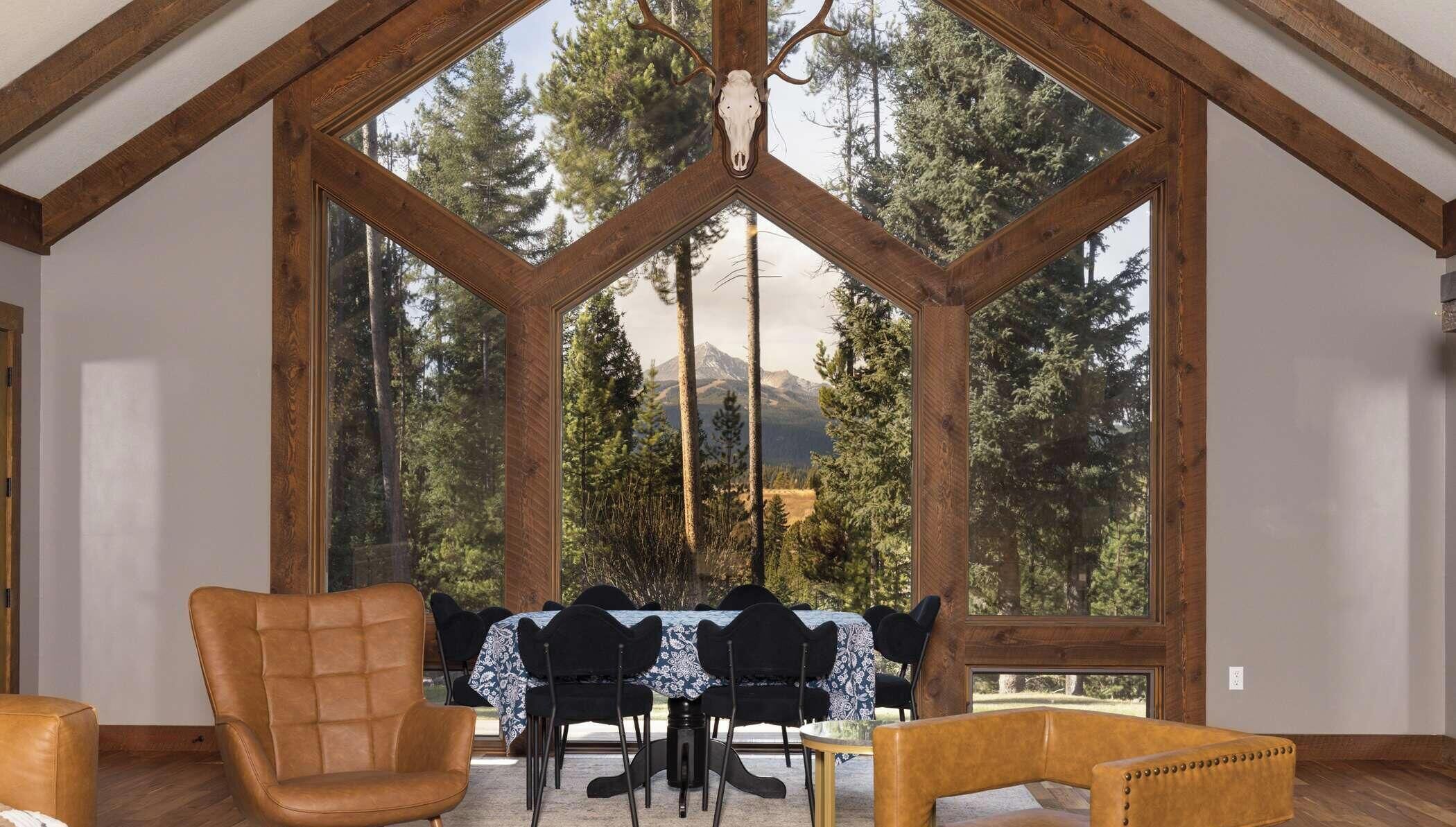 Ein Zimmer mit einem großen Fenster mit Holzrahmen, einem Esstisch mit Stühlen und Blick auf einen Wald und Berge.