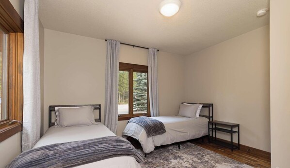 Quarto com duas camas de solteiro, mesa de cabeceira, janela com cortinas e luminária no teto.
