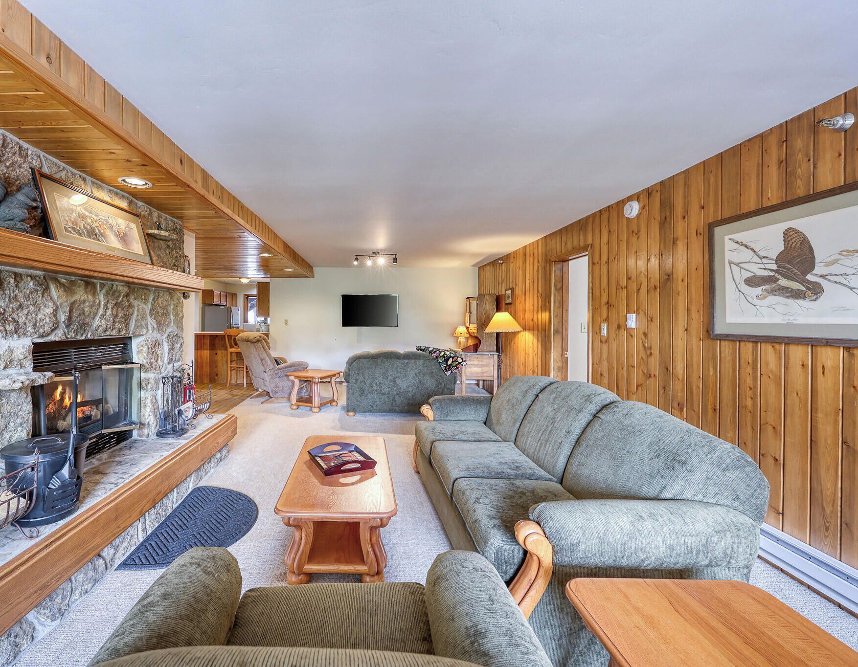 A cozy living room with a stone fireplace, wooden walls, a grey sofa, and a television mounted on the wall.