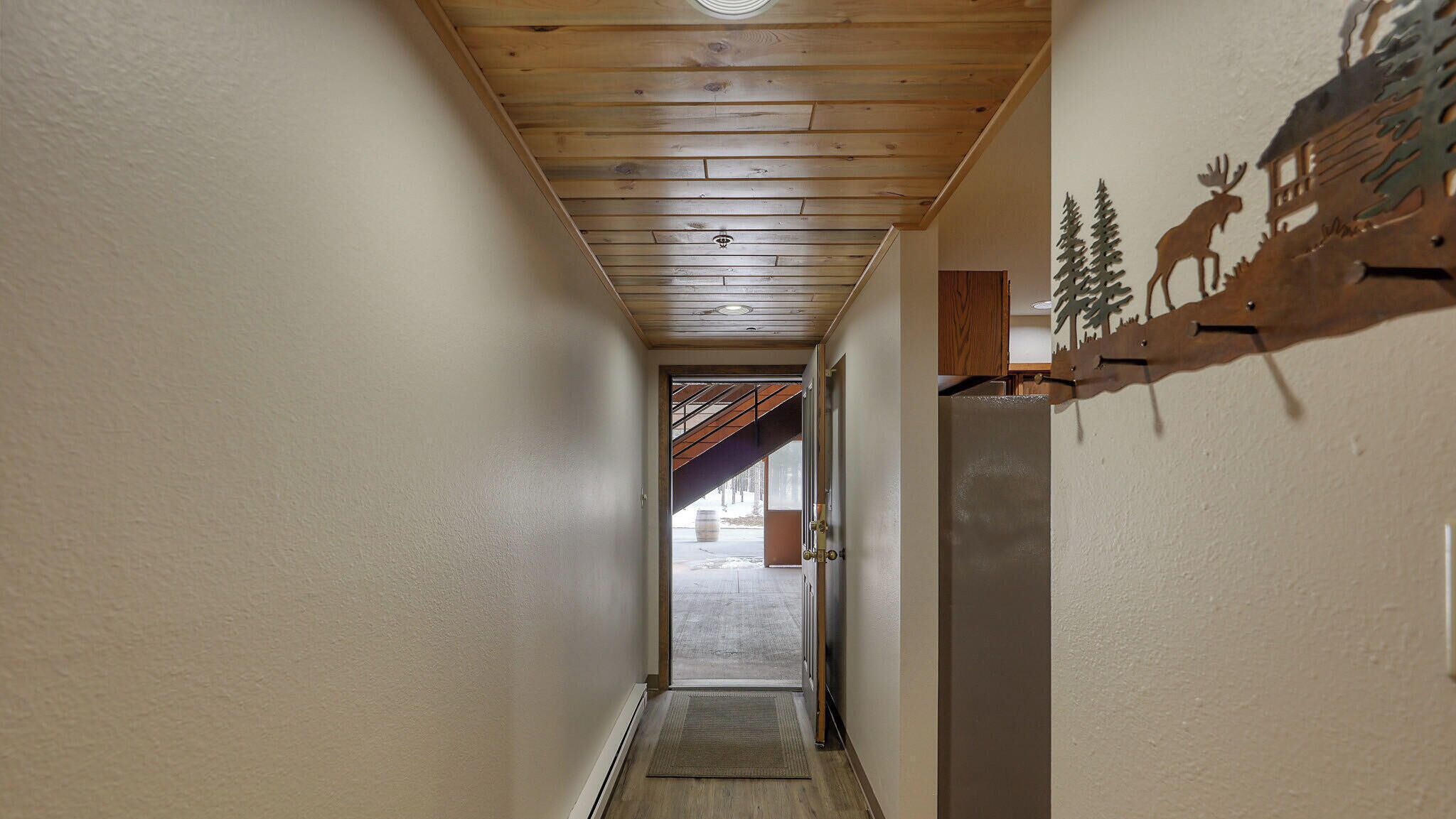 A narrow hallway with wooden ceiling beams, a door leading to an outdoor area, and a wall-mounted decoration of a moose and trees.