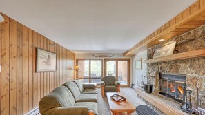 A cozy living room with a stone fireplace, wooden walls, and a large window. - Mountain Village Gem! Gorgeous Skycrest Condo (Big Sky)