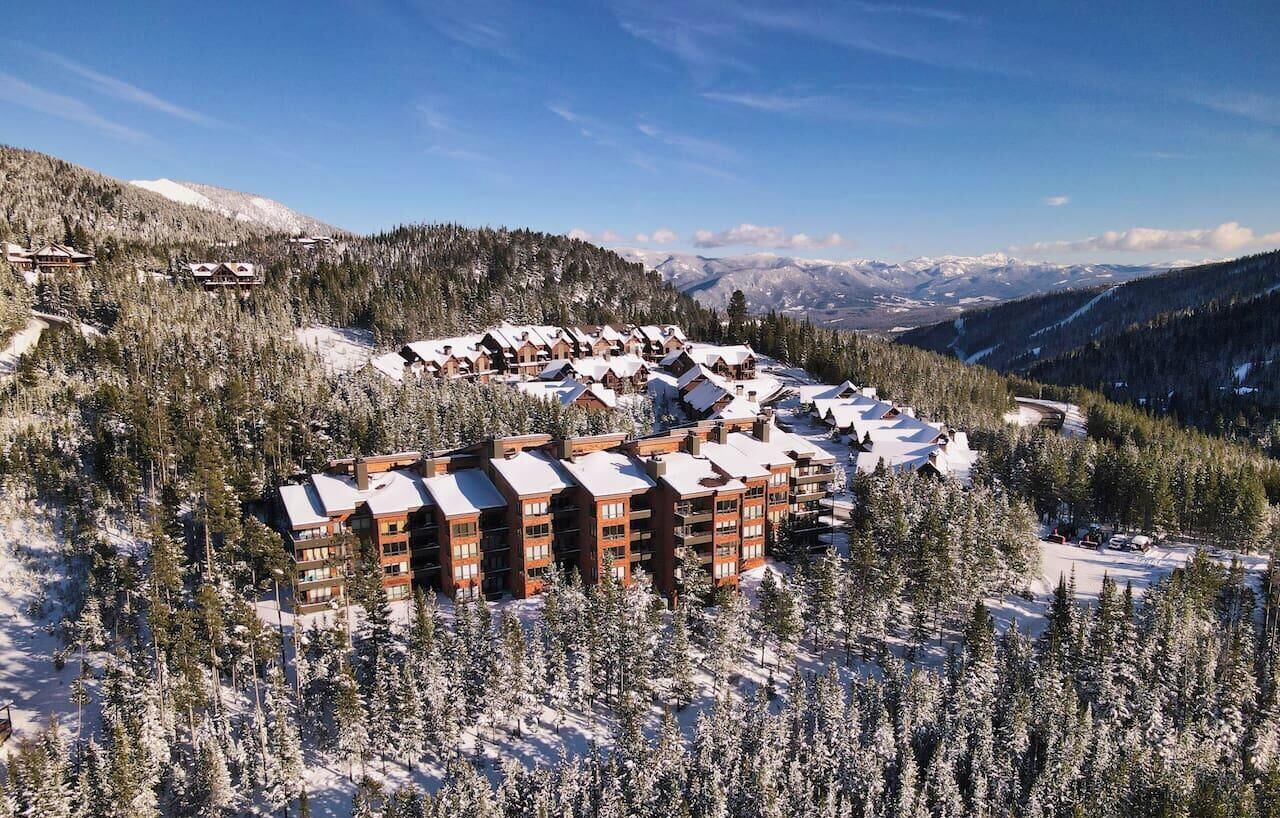 A snow-covered lodge with multiple buildings, surrounded by dense forest and mountains in the background.