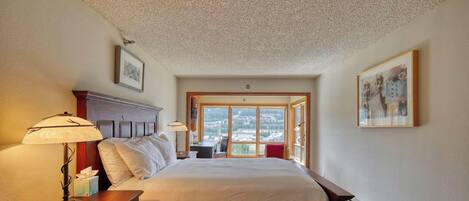 A hotel room with a large bed, a nightstand with a lamp, a window with a view of the city, and artwork on the walls.